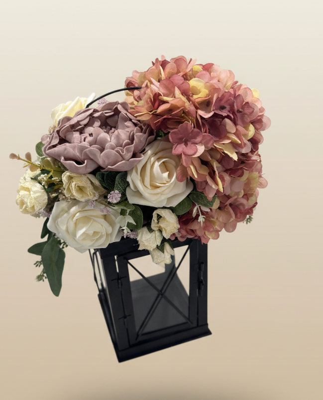 Artificial flower arrangement with pink and white roses, purple and yellow hydrangeas, and greenery in a black lantern-shaped container against a plain background.