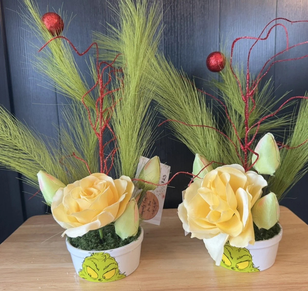 Two decorative plant arrangements in white pots with a Grinch face design, featuring yellow roses, green foliage, red curly accents, and green grass-like leaves, placed on a wooden surface against a blue background.
