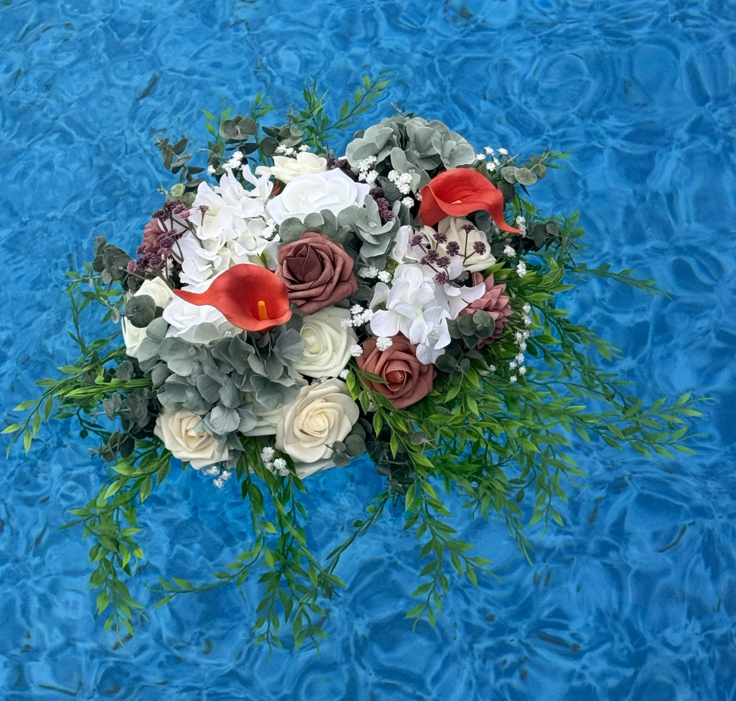 A floral arrangement with white, gray, maroon, and red flowers, surrounded by green foliage, floating on a blue water background.