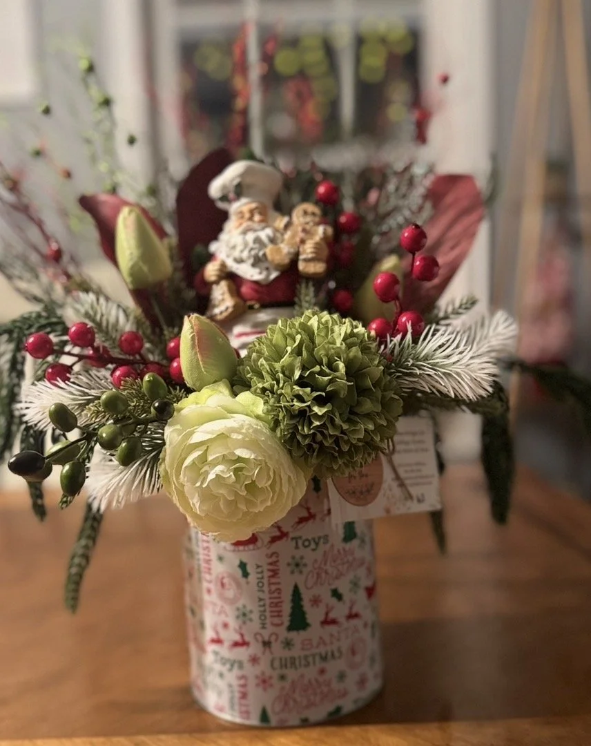 A festive Christmas flower arrangement in a decorated container featuring Santa figurines, red berries, green foliage, and white accents, placed on a wooden surface.