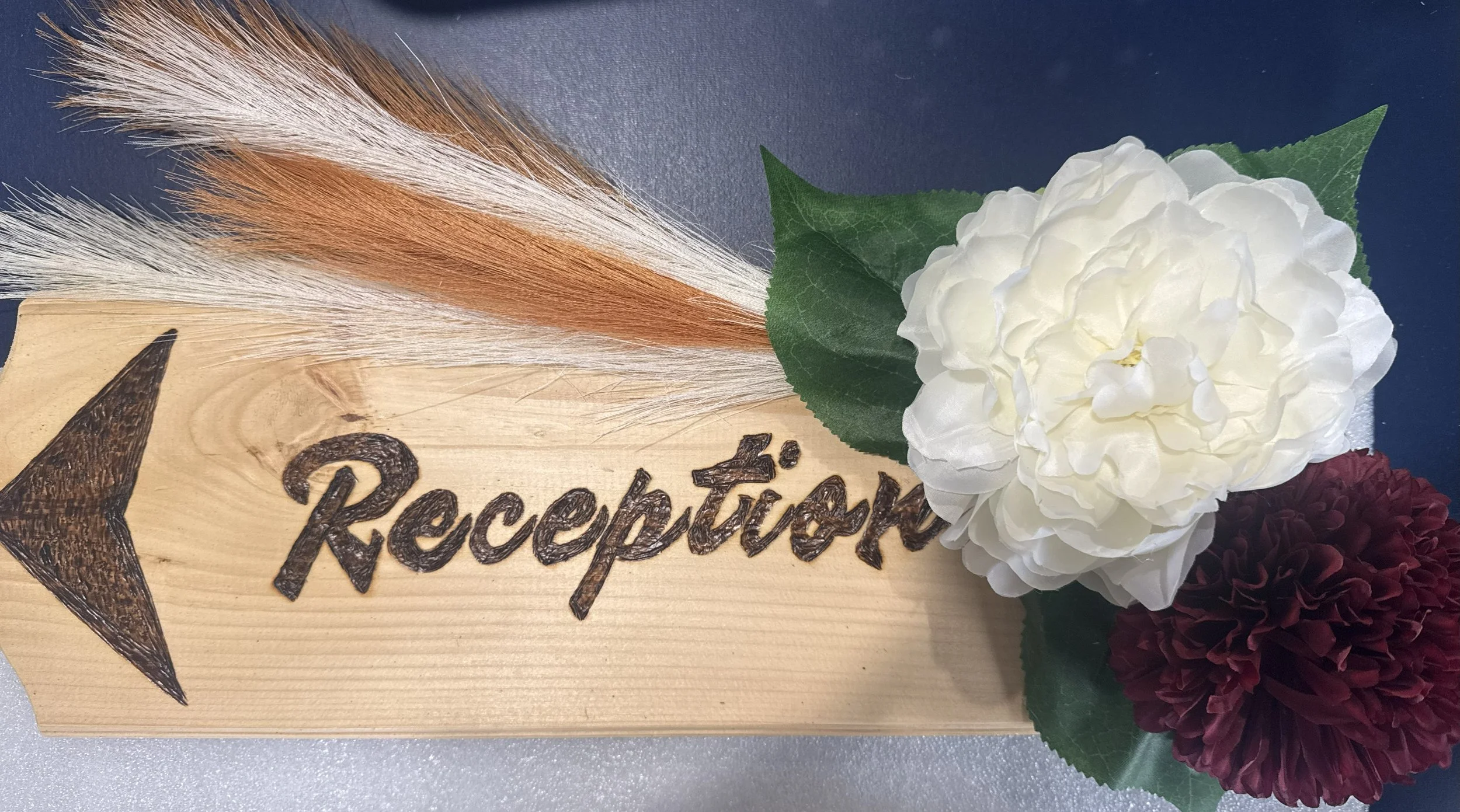 A wooden reception sign with an arrow and the word 'Reception' burned into the wood. There are decorative flowers, including white and dark red blooms, and dried grasses placed on the right side of the sign.