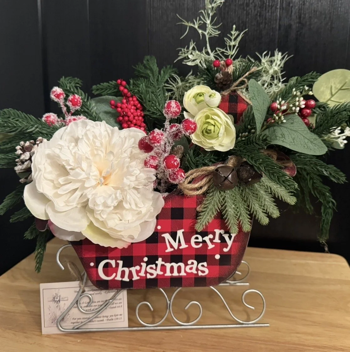 A Christmas floral arrangement in a red and black plaid sleigh-shaped container with the text 'Merry Christmas'. The arrangement includes white flowers, red berries, pinecones, and green foliage, set on a wooden surface.