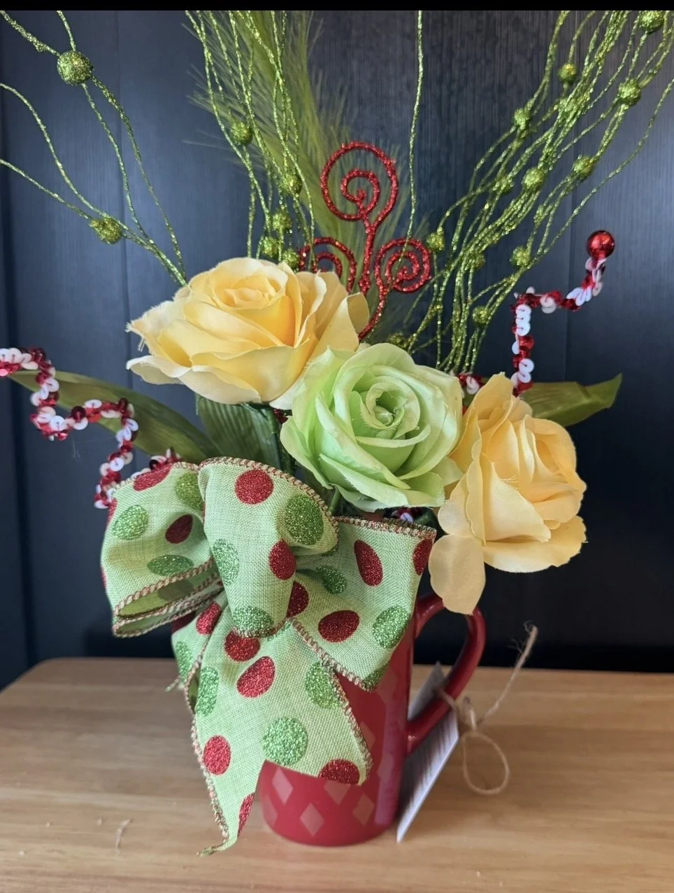 Decorative floral arrangement in a red mug, with yellow and green roses, glittery green sprigs, a polka-dot ribbon bow, and red and white beaded accents, placed on a wooden surface.
