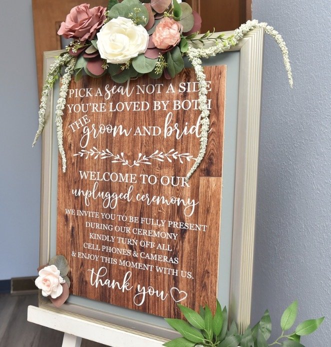 Wooden wedding sign decorated with pink and white flowers and greenery, containing wedding instructions and a thank you message.