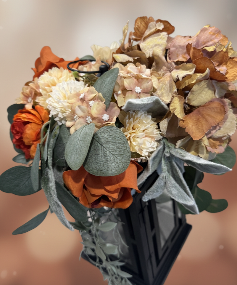 A bouquet of artificial flowers in shades of orange, cream, and brown with green and gray leaves arranged in a black geometric vase.