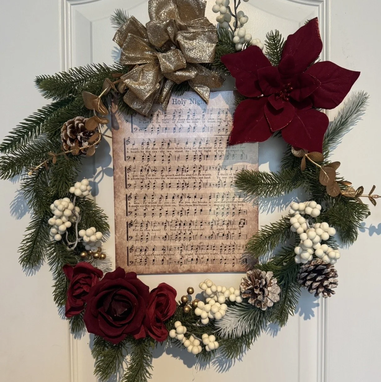 Christmas wreath decorated with pine branches, red roses, pinecones, white berries, gold leaves, a large gold bow, a large red poinsettia, and a sheet of music titled 'Holy Night' at the center.