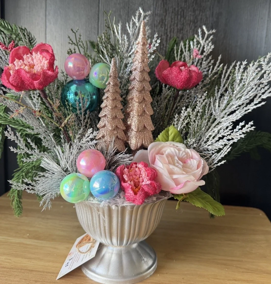 A festive floral arrangement with pink, peach, and green flowers, decorated with iridescent and metallic Christmas ornaments, silver pine branches, and glittery gold conical tree decorations in a silver vase.