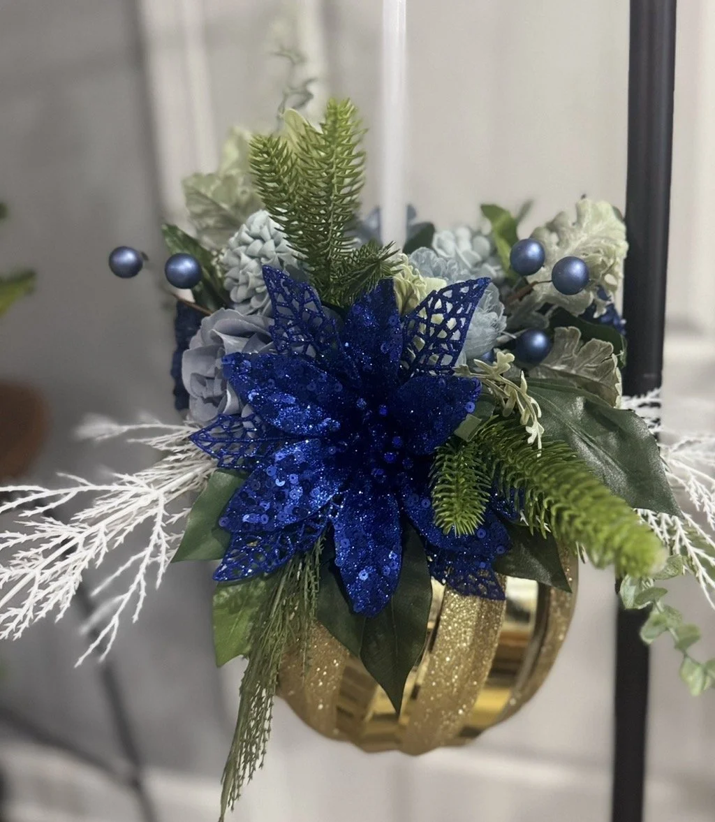 Decorative Christmas floral arrangement with a large blue glittery poinsettia, green pine branches, white and blue berries, and white and green foliage, in a gold and black vase.