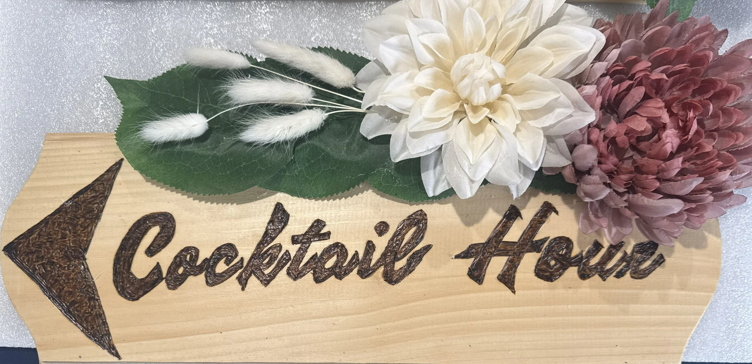 Decorative wooden sign with the words 'Cocktail Hour' written on it, adorned with artificial white and pink flowers and green leaves.