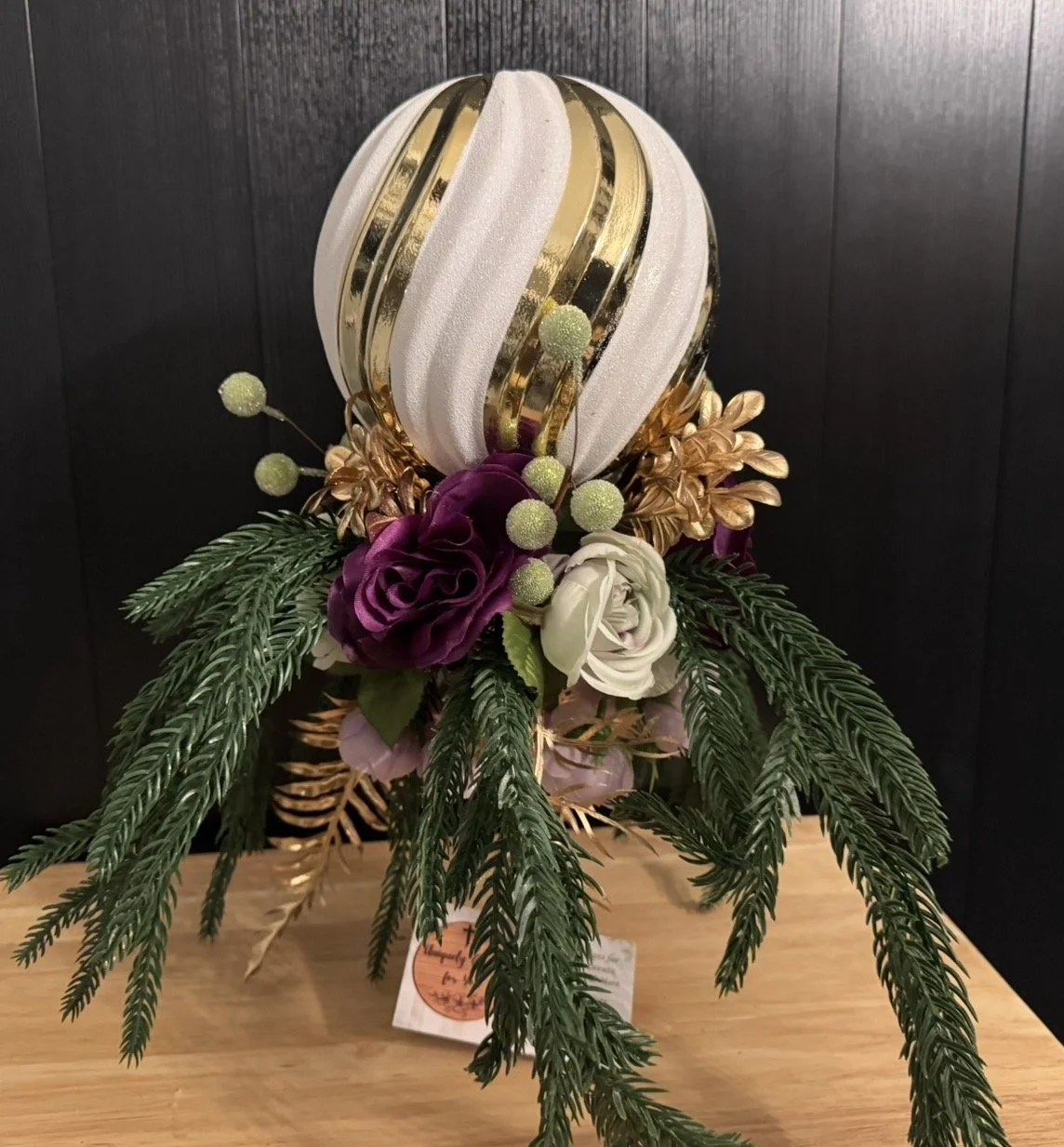 Decorative Christmas floral arrangement with a large ornament, purple and white roses, gold leaves, and pine branches.