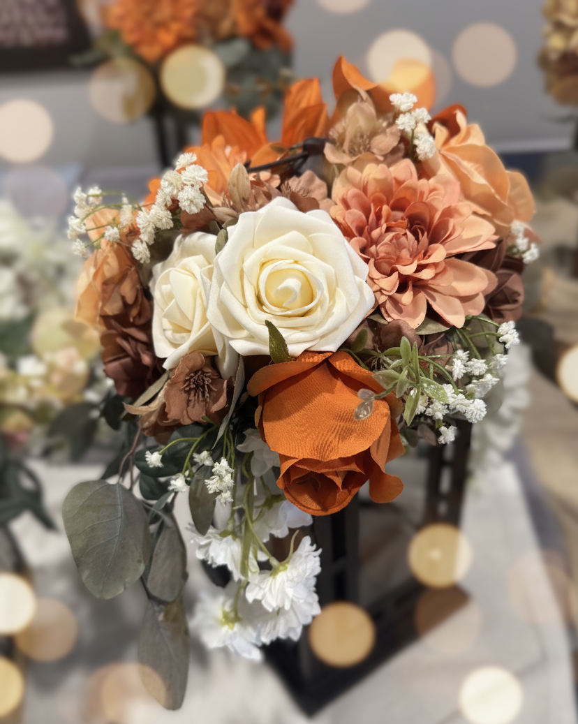 Artificial floral arrangement with white roses, orange and brown flowers, small white filler flowers, and green leaves, displayed on a black stand.