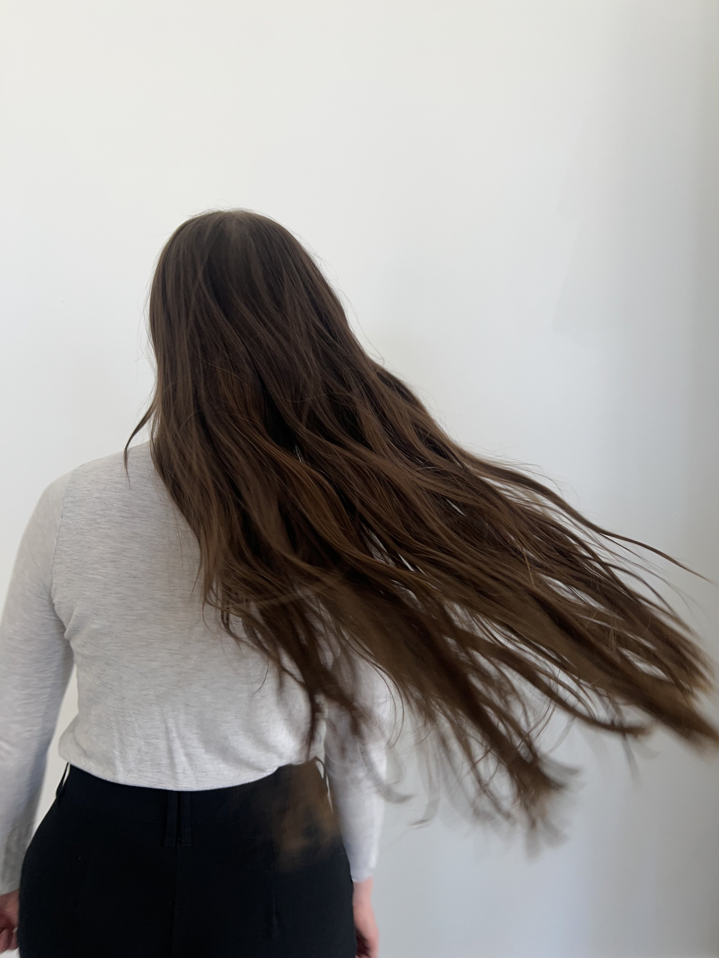 Person with long brown hair turning, wearing a light gray sweater, against a white background.