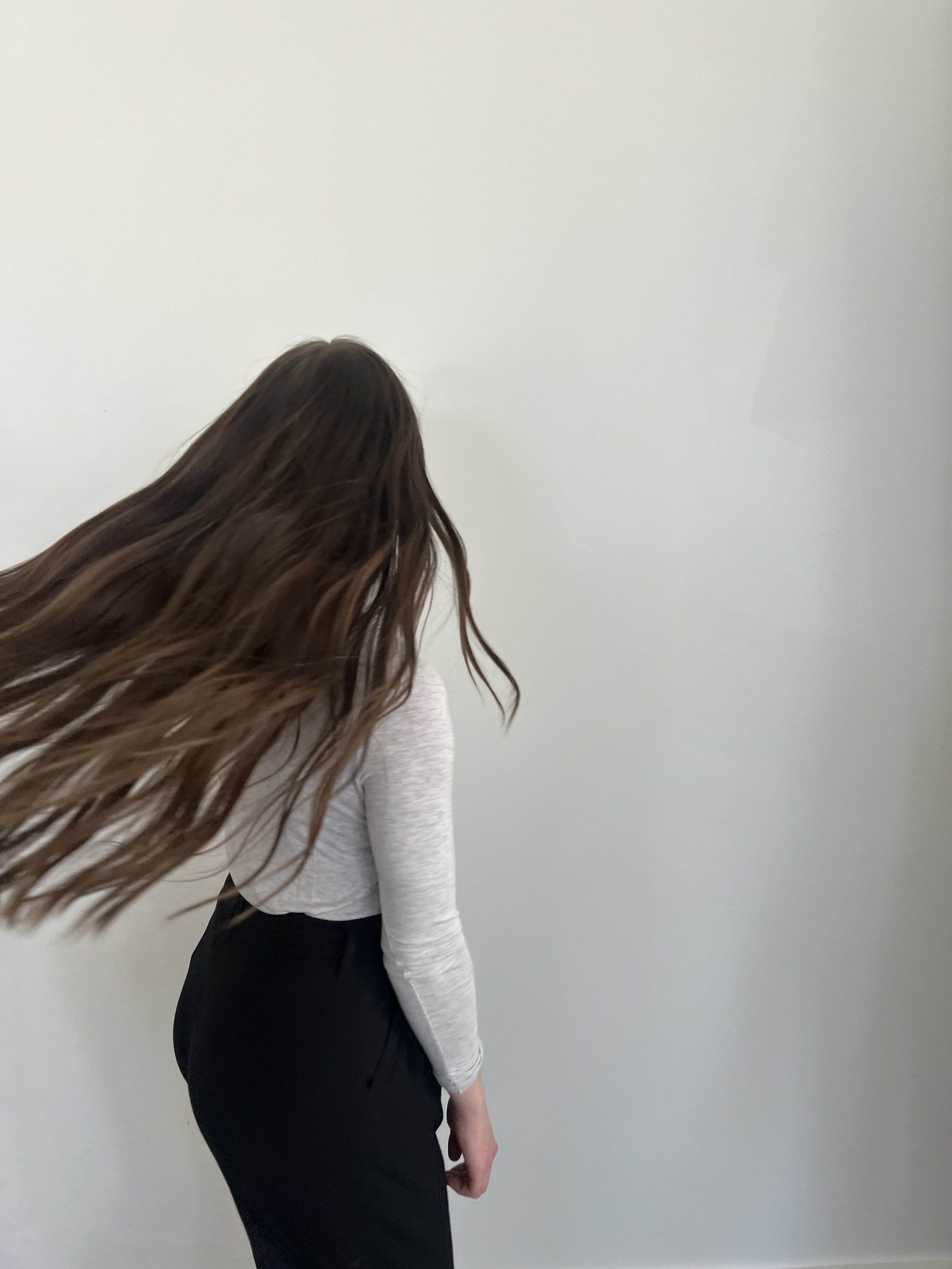 Person with long hair and motion blur, wearing a gray top and black skirt, turning away from the camera against a plain white background.