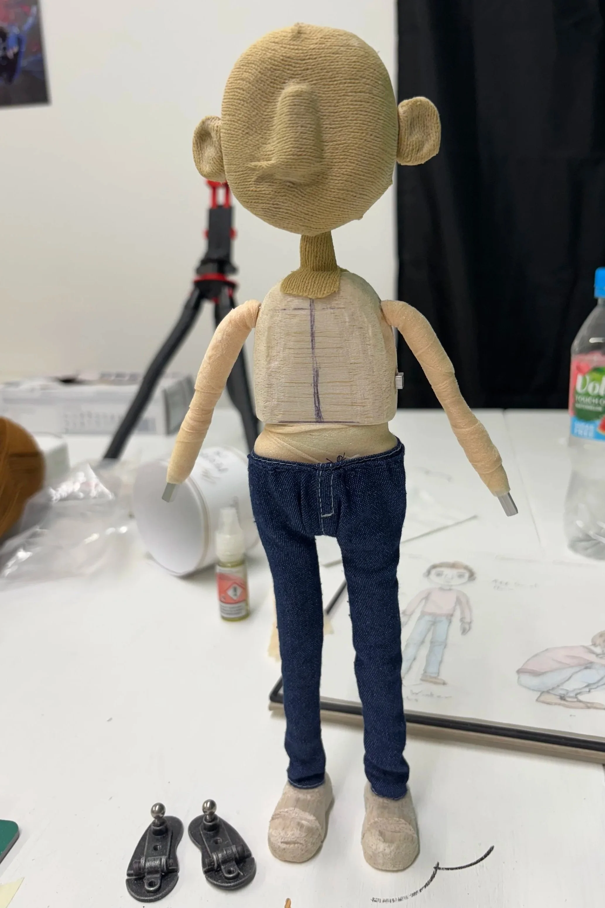 A handmade fabric and wood doll with a large head, dressed in blue jeans and beige shoes, standing on a table surrounded by art supplies and sketches.