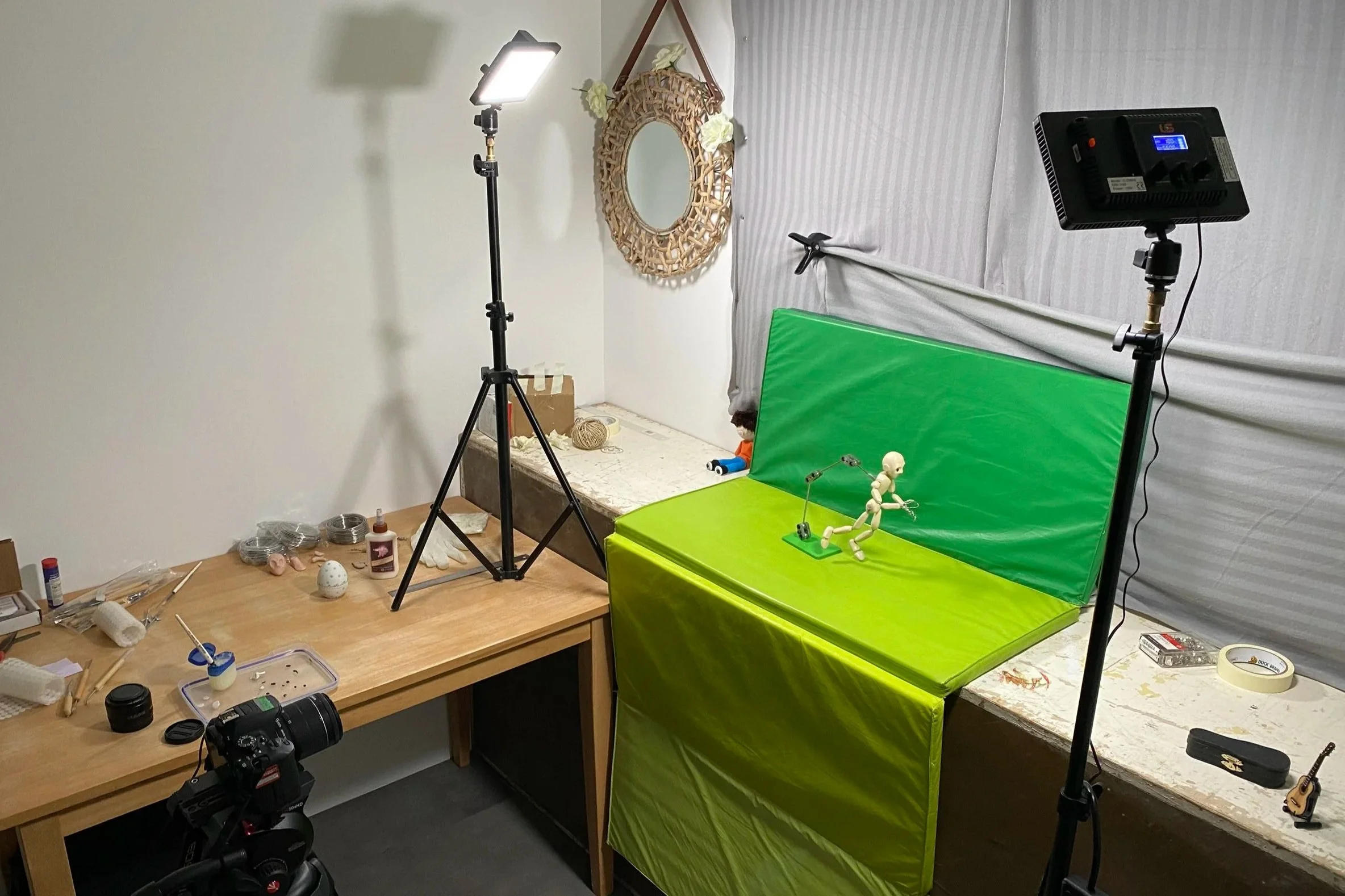 Inside a small room, a camera setup with lighting equipment pointing at a green screen backdrop. There is a desk with craft supplies and a small doll mannequin in a running pose on a green platform, as part of a setup for video or photo recording.