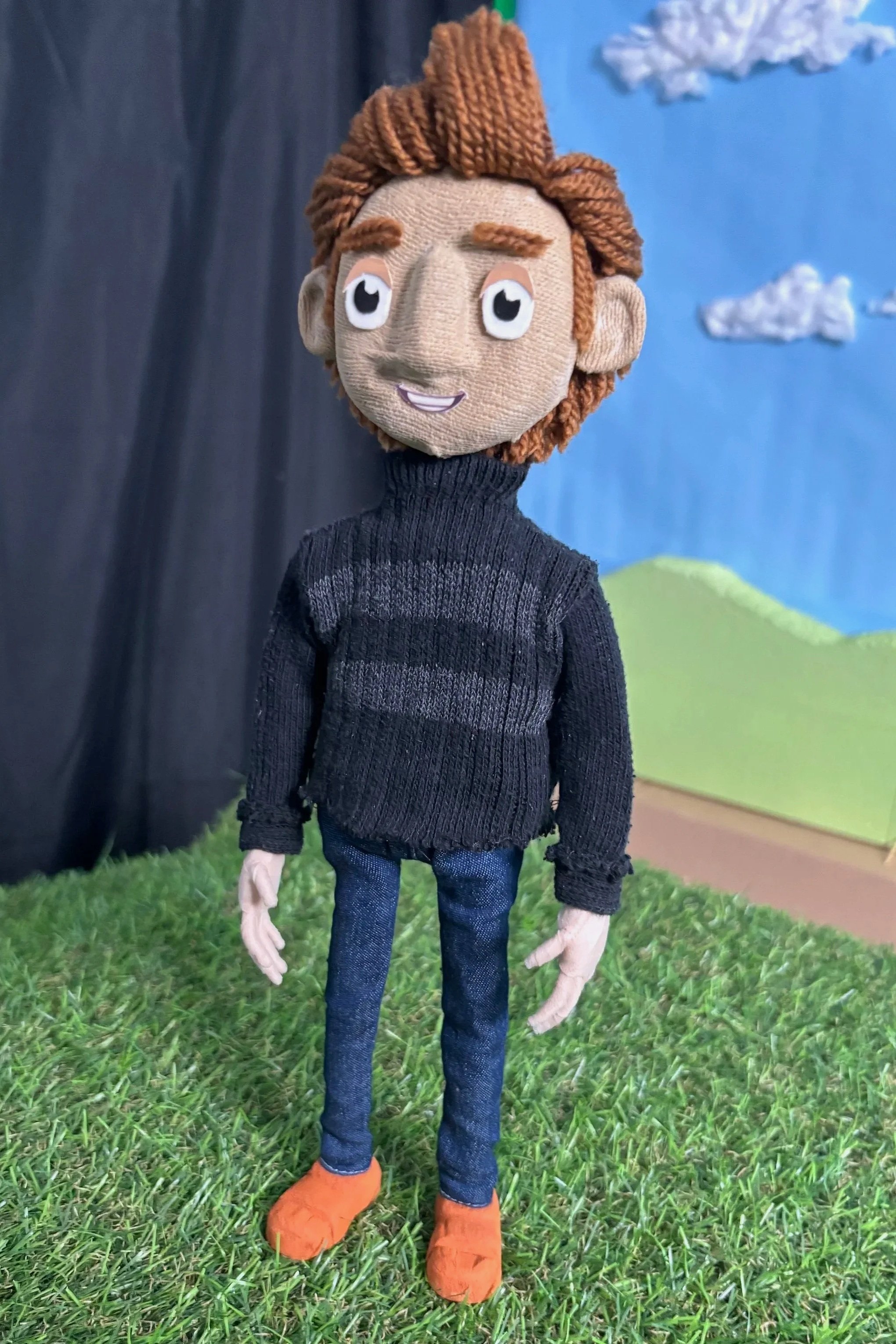 A puppet character with brown yarn hair, a beige face, and expressive eyes. Wearing a black turtleneck sweater, blue jeans, and orange shoes, standing on a grassy surface with a backdrop of a blue sky with clouds and green hills.