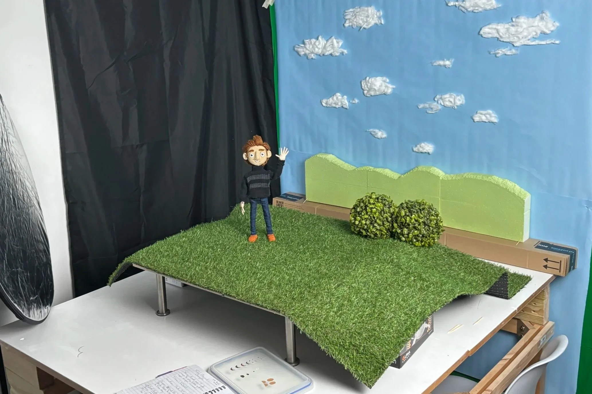 Miniature scene with a puppet figure waving, set on a platform covered with artificial grass, with a background mural of clouds and hills, and small trees made of foam or similar materials.
