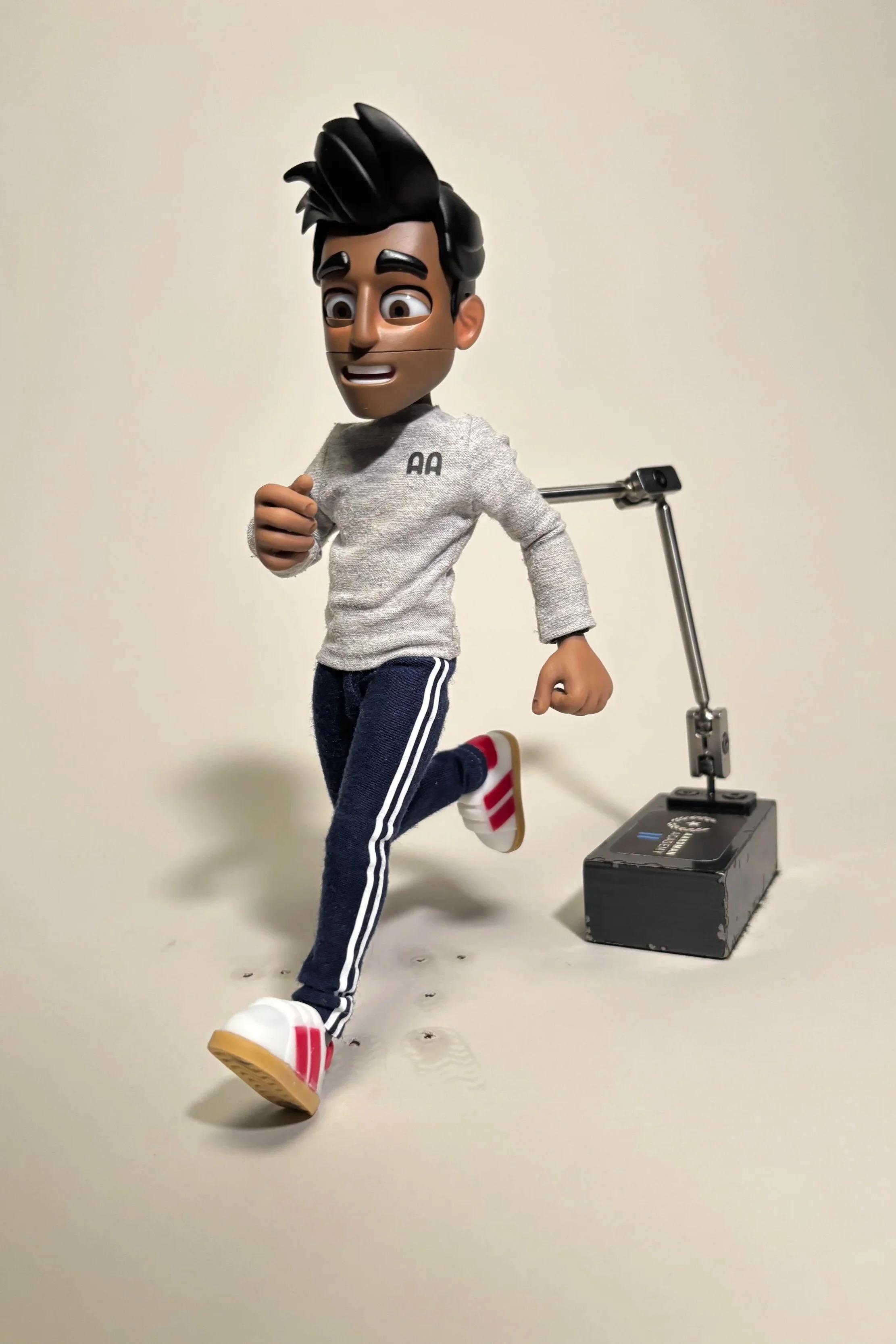 A running animated character with a stylized human appearance, wearing a gray sweatshirt, navy blue track pants with white stripes, and white sneakers with red accents, on a plain light-colored background.