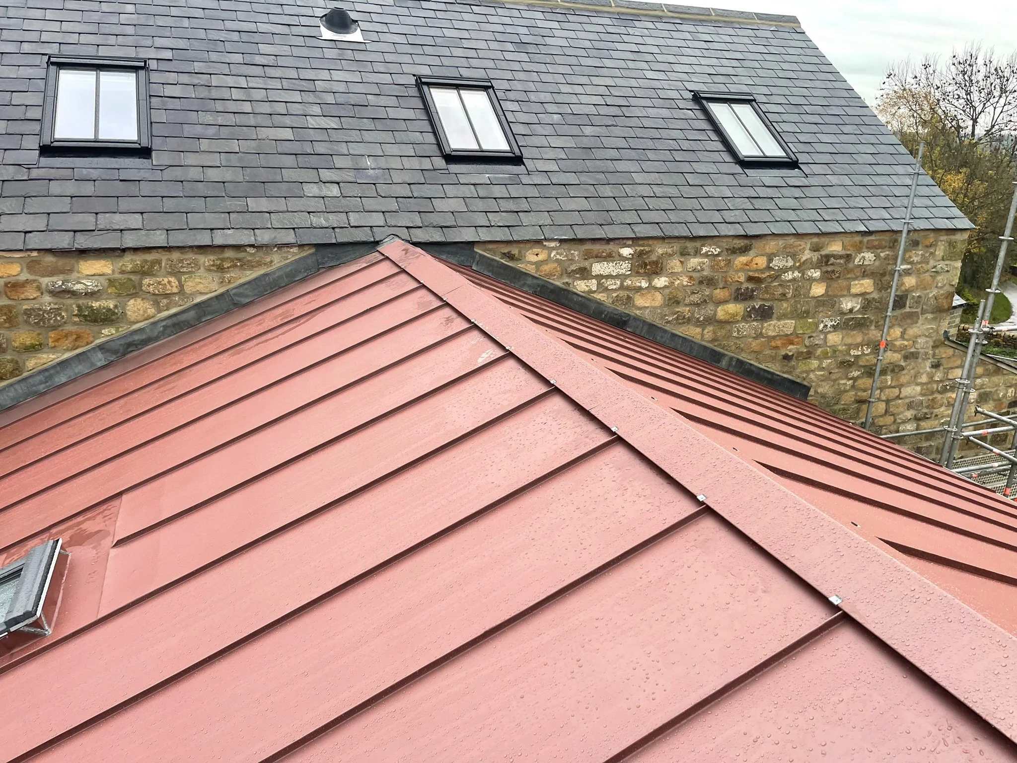 A red metal roof with raindrops visible, in front of a stone building with a slate roof featuring two skylights.