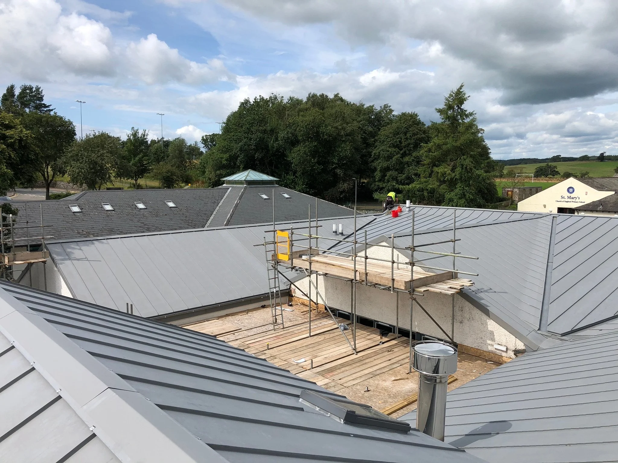 Construction site with scaffolding and new metal roofing, surrounded by trees and sky.