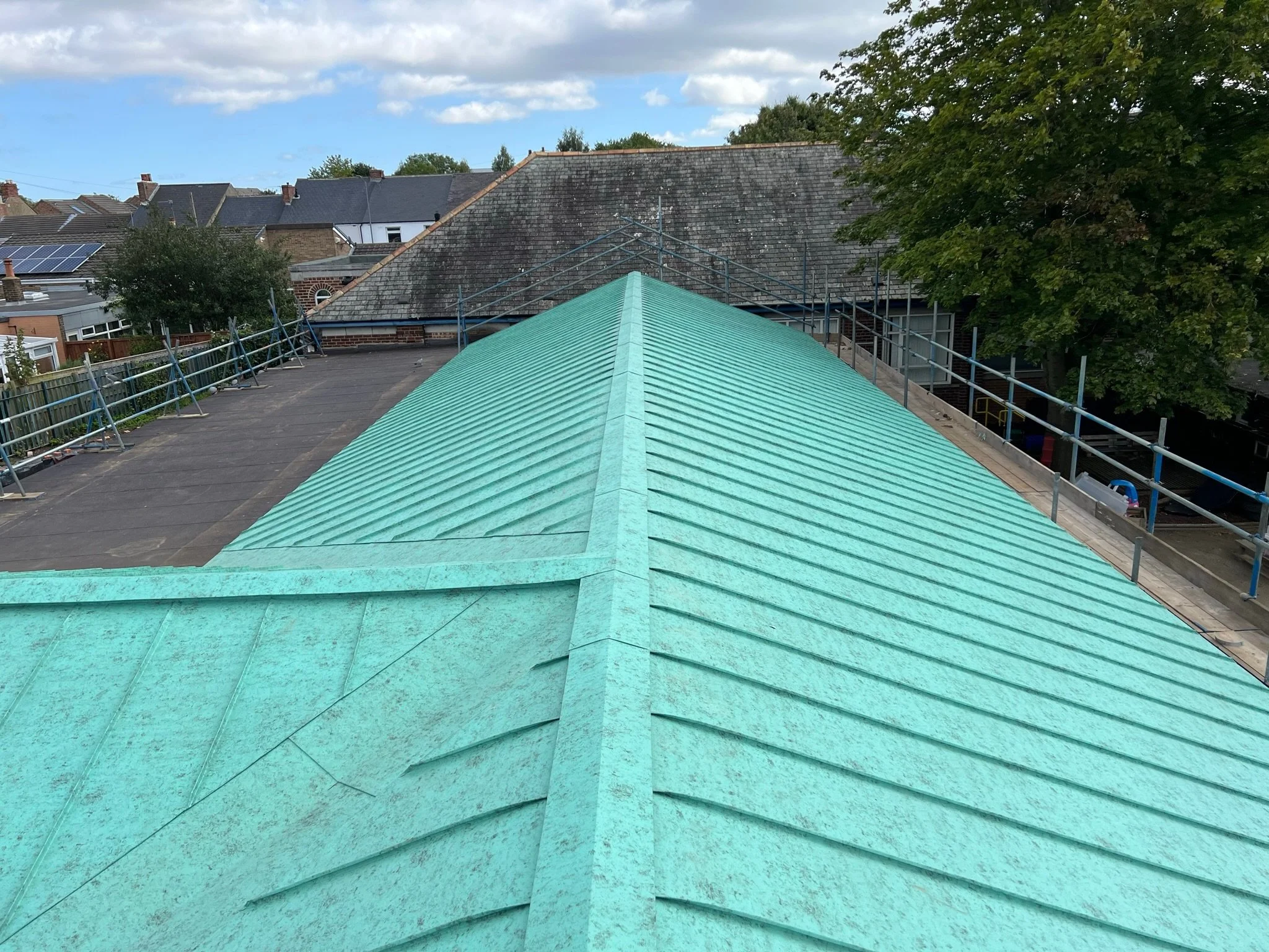 Green metal roof with scaffolding on sides, trees and houses in background.