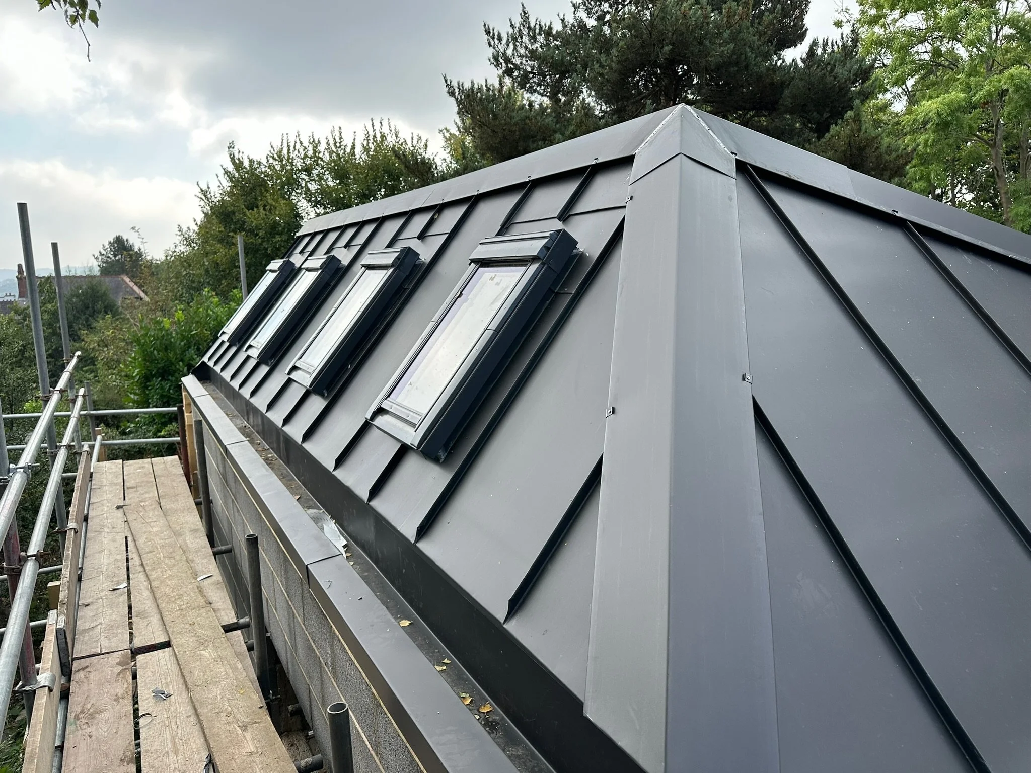 Metal roof with multiple skylights on a construction site, surrounded by scaffolding, with trees in the background.