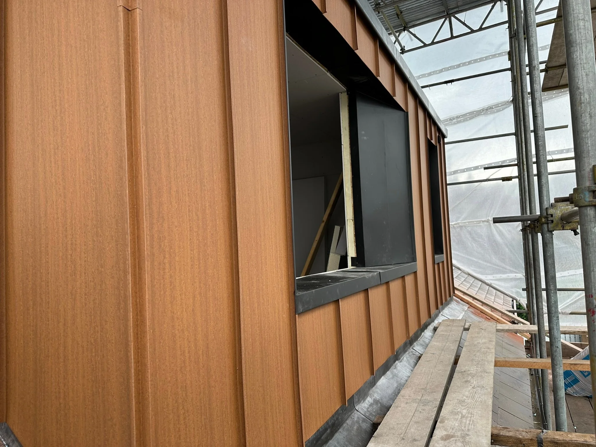 Close-up of building under construction with orange metal siding and scaffolding, showing window opening.