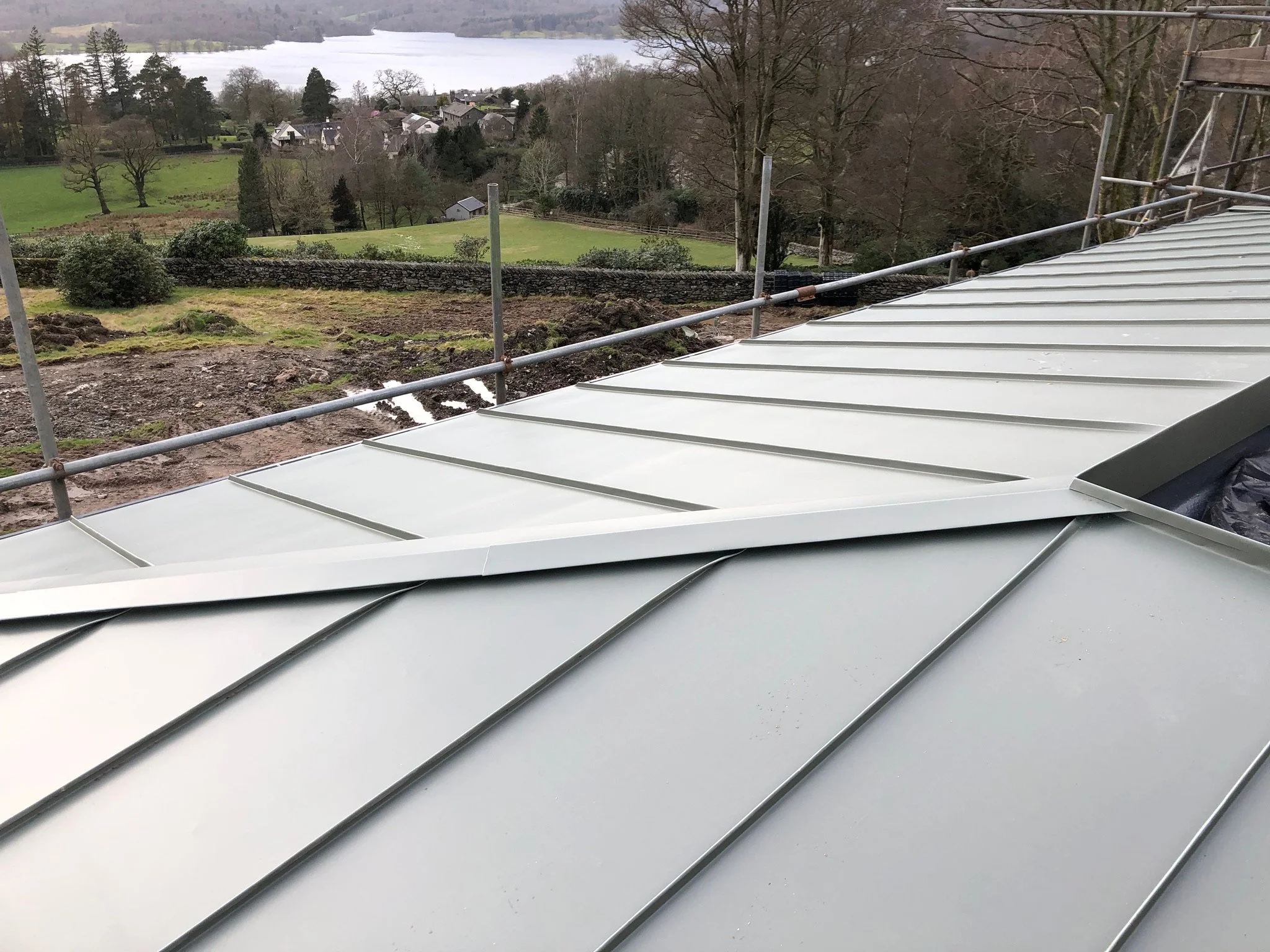 Green metal roof with a scenic countryside background, including trees, fields, and a lake. Scaffolding is visible beside the roof.