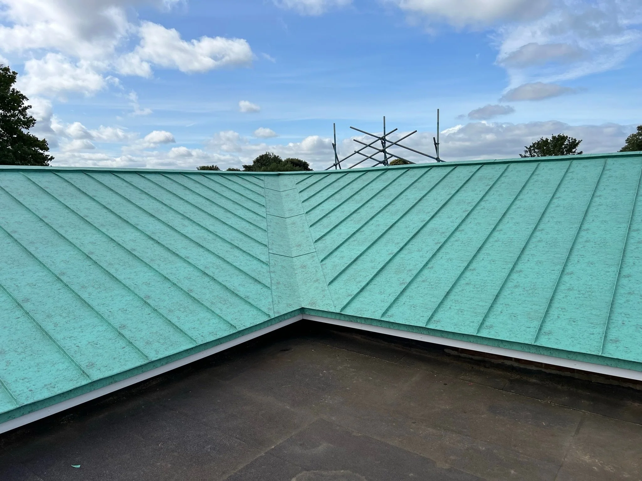 A green metal roof with a standing seam design against a blue sky with clouds and scaffolding in the background.