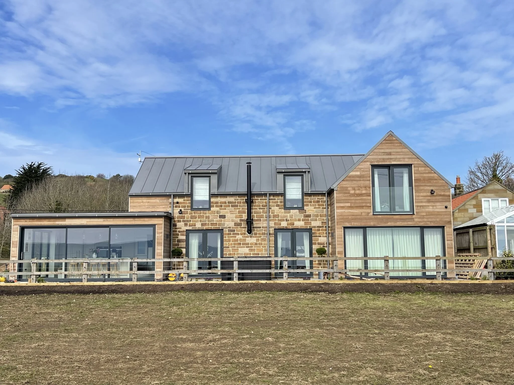 Modern house with brick and wood exterior, large windows, and a sloped metal roof, set in a rural landscape with a clear sky in the background.