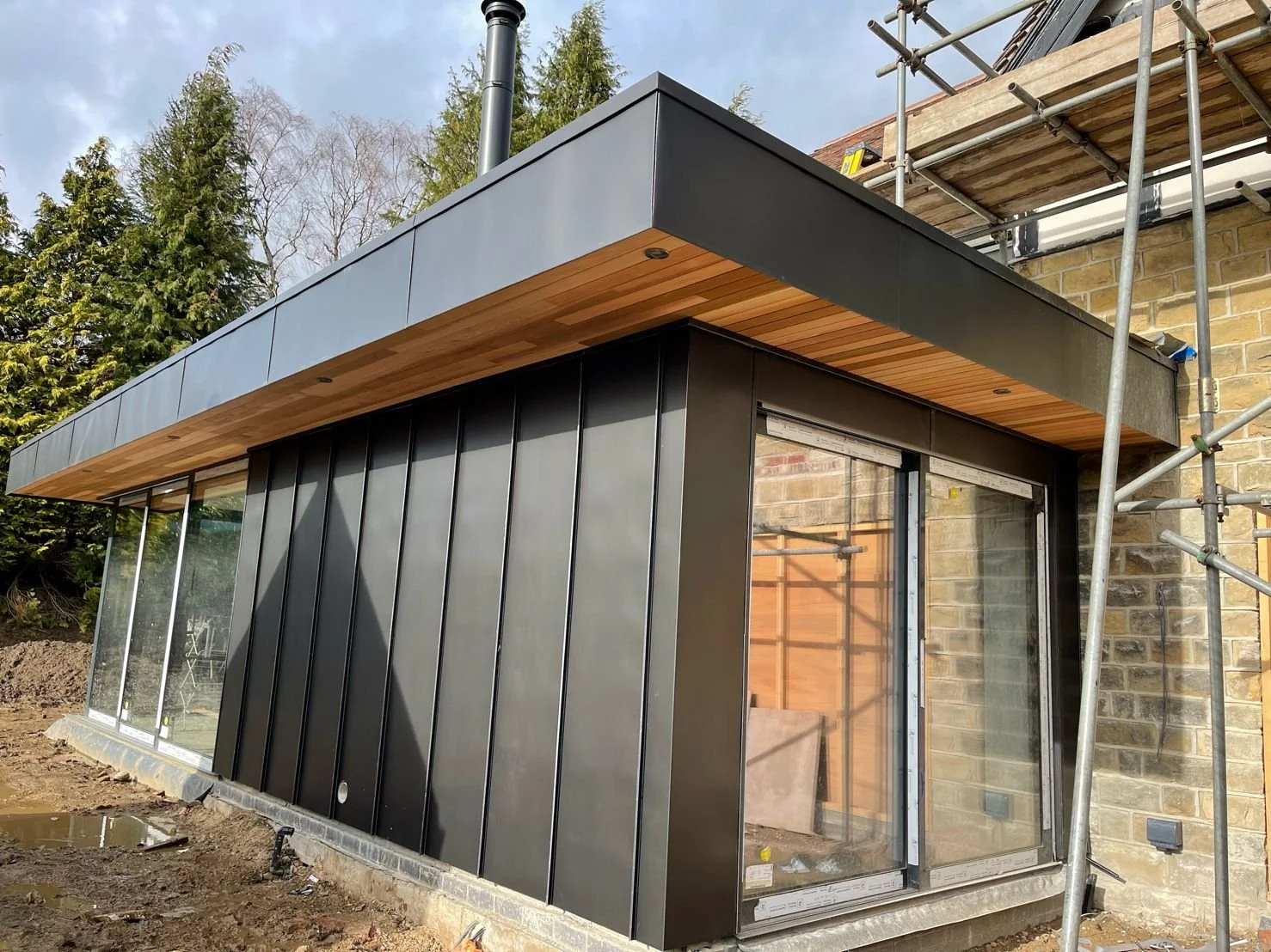 Modern building extension under construction with glass doors, black metal cladding, and wooden accents, surrounded by scaffolding and trees.
