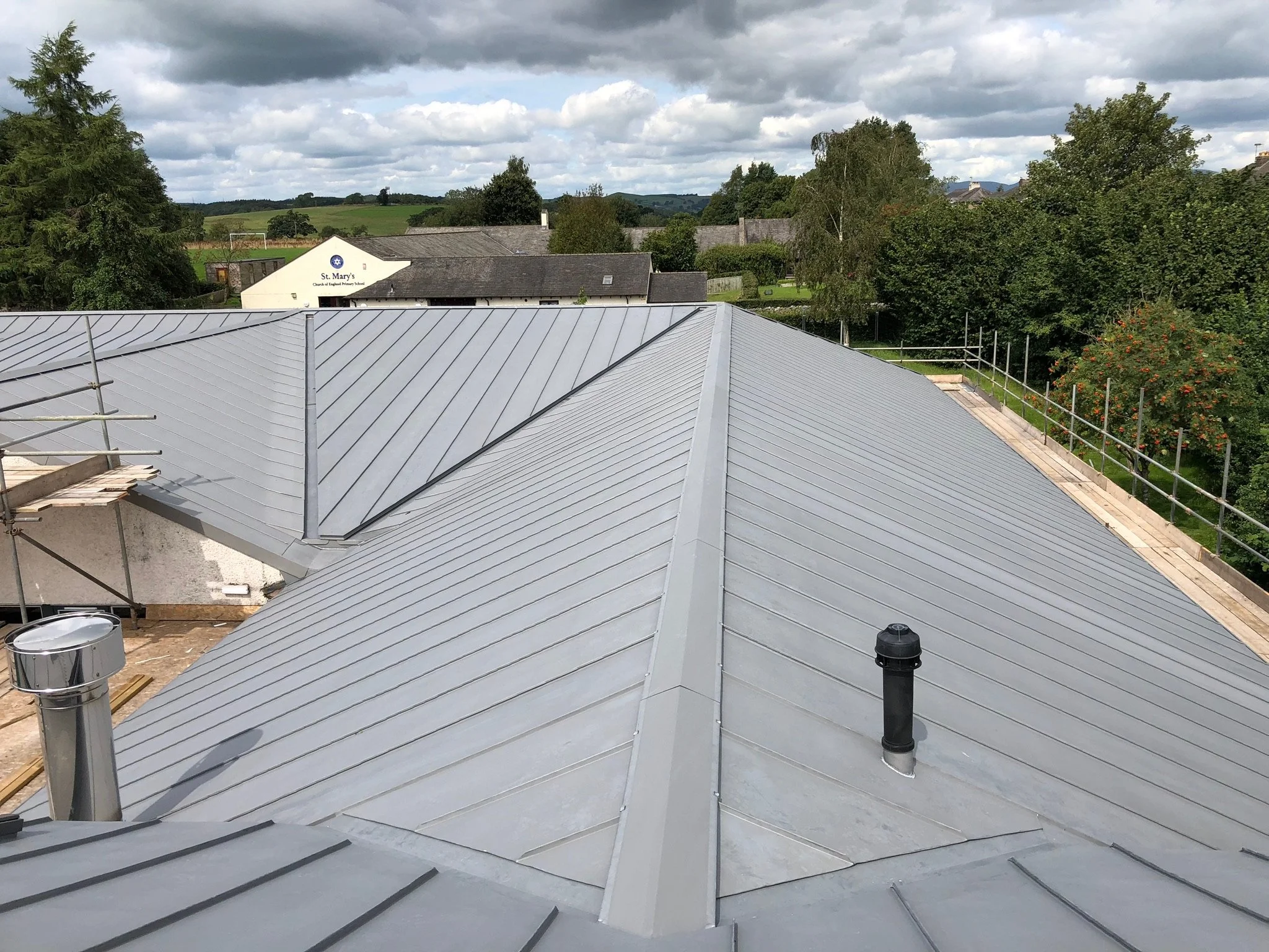 View of a metal roof with standing seam panels and surrounding scaffolding. In the background, green trees and a building with a sign reading 'St. Mary's' are visible under a cloudy sky.