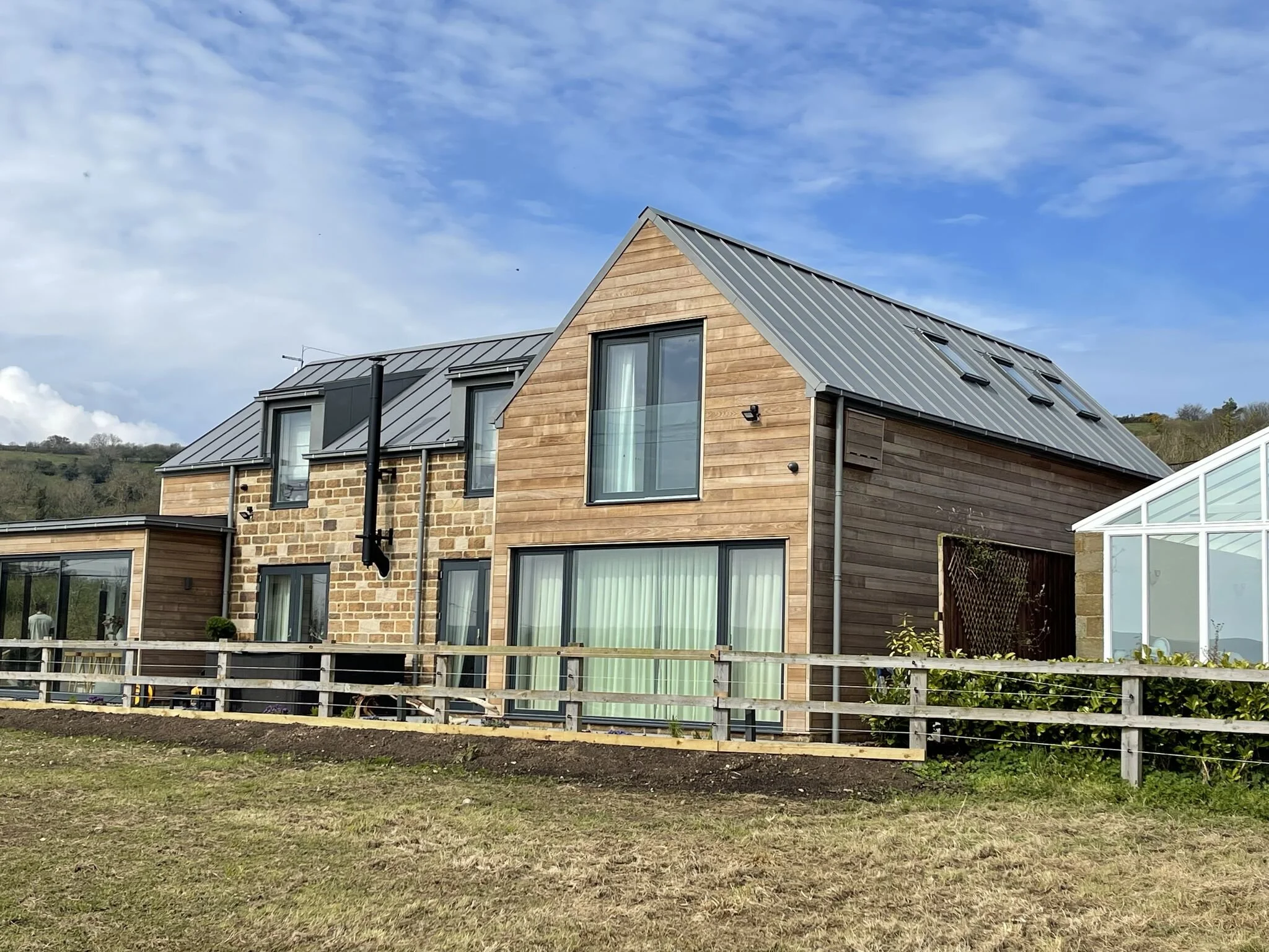 Modern house with wood and stone exterior, large windows, metal roof, and glass extension.