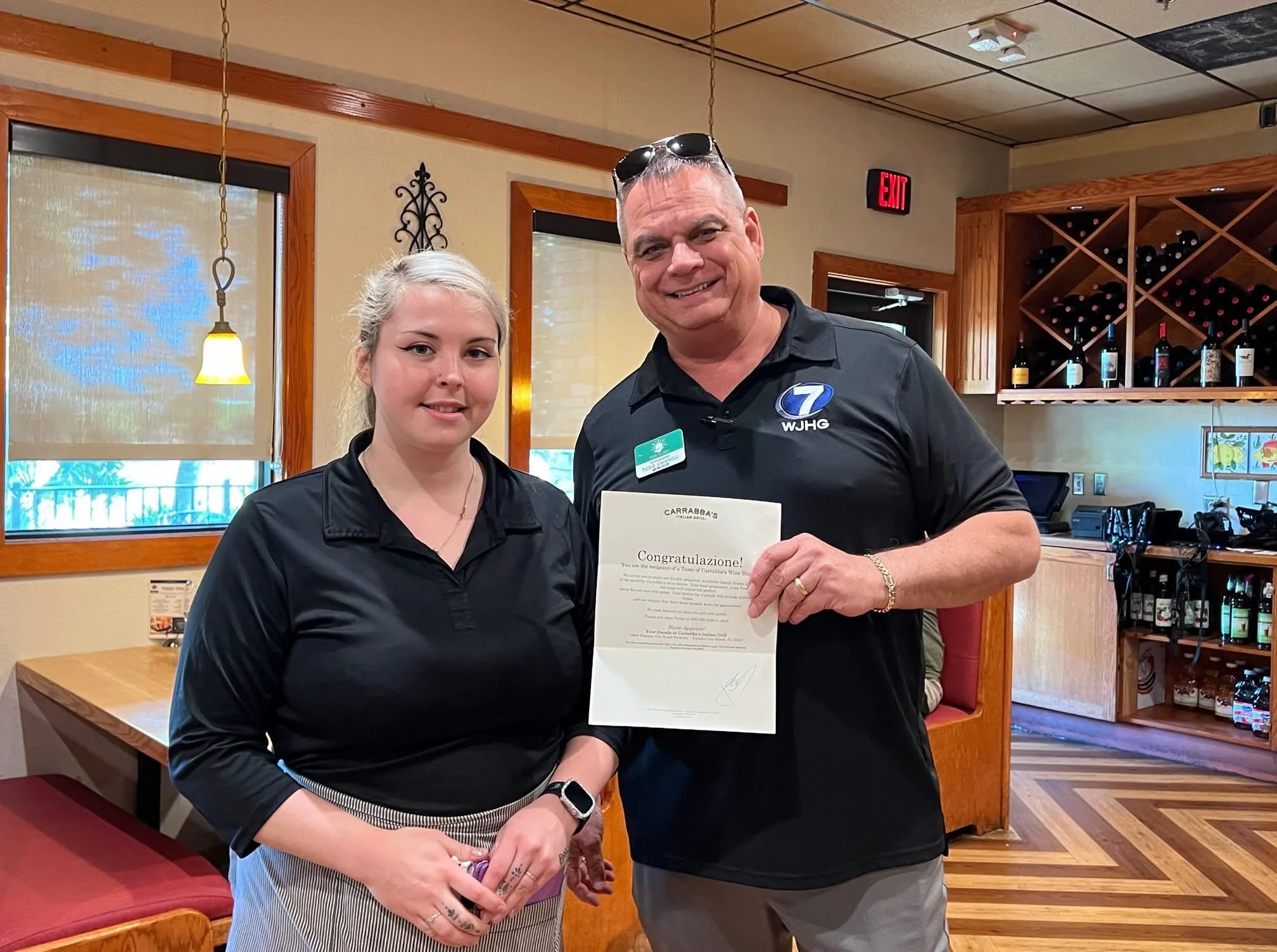 A man and a woman standing side by side inside a restaurant, holding a certificate of recognition. The man is smiling and wearing a black polo shirt with the logo 'WJHG' and sunglasses on his head. The woman has blonde hair tied back, wearing a black