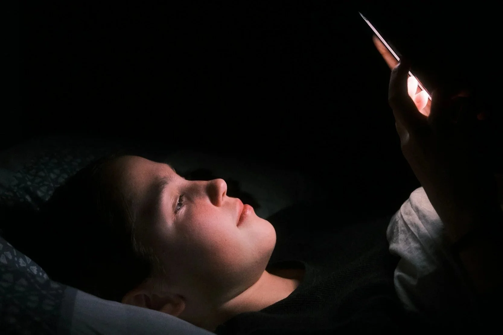Person lying in bed in darkness, illuminated by the light of a smartphone they are holding
