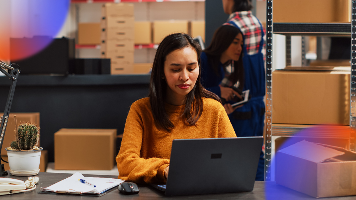Warehouse employee working on a laptop while inventory and shipping operations take place in the background, representing e-commerce logistics, inventory management, and order fulfillment.