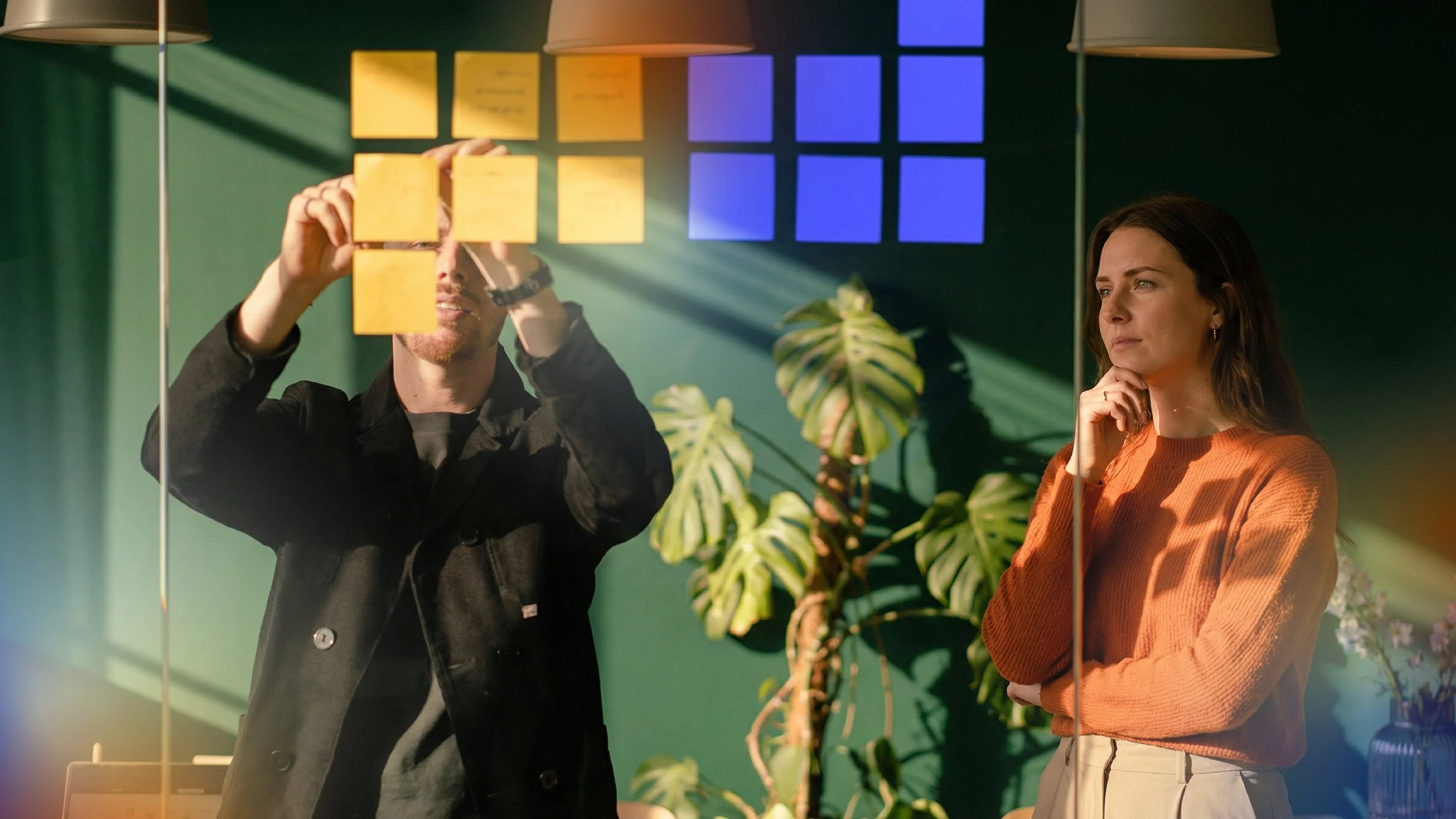 Dois profissionais analisando notas e gráficos de desempenho em um quadro de vidro, representando trabalho em equipe, planejamento estratégico e decisões de mídia baseadas em dados.