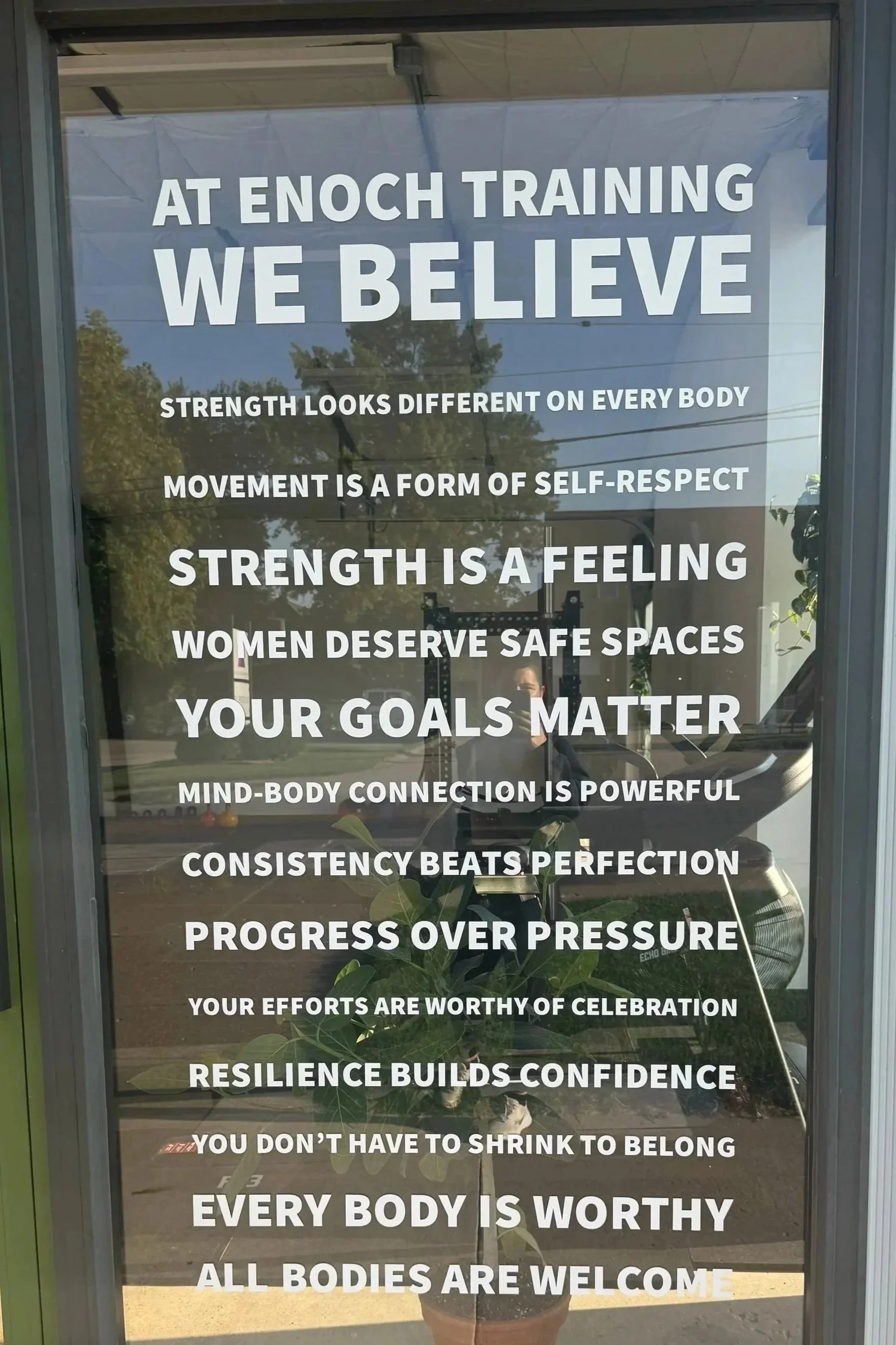Motivational statements about strength, self-respect, goals, resilience, and inclusivity displayed on a glass door at Enoch Training, with reflections of trees, a person, and exercise equipment outside.