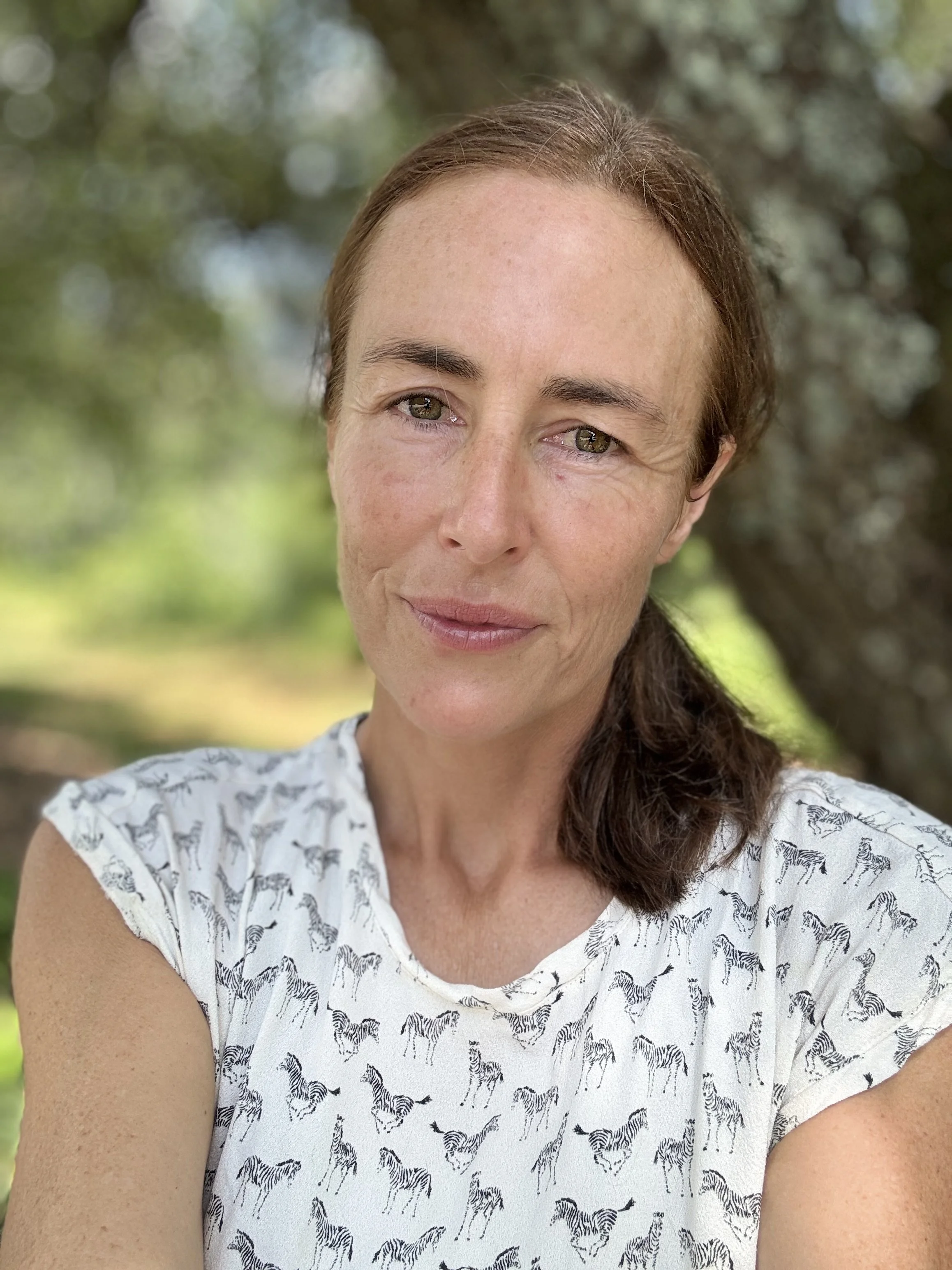 A woman with brown hair, light skin, and green eyes taking a selfie outdoors with a blurred green and brown background, wearing a white shirt with zebra print.
