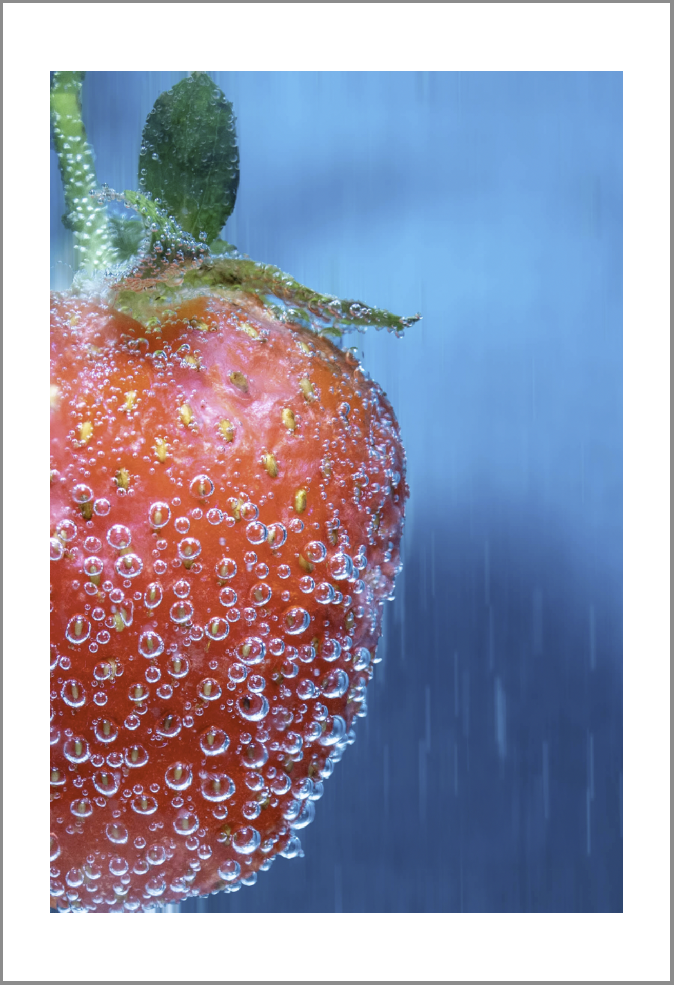 Close-up of a red strawberry submerged in water with bubbles on its surface and green leaves on top, against a blue background.