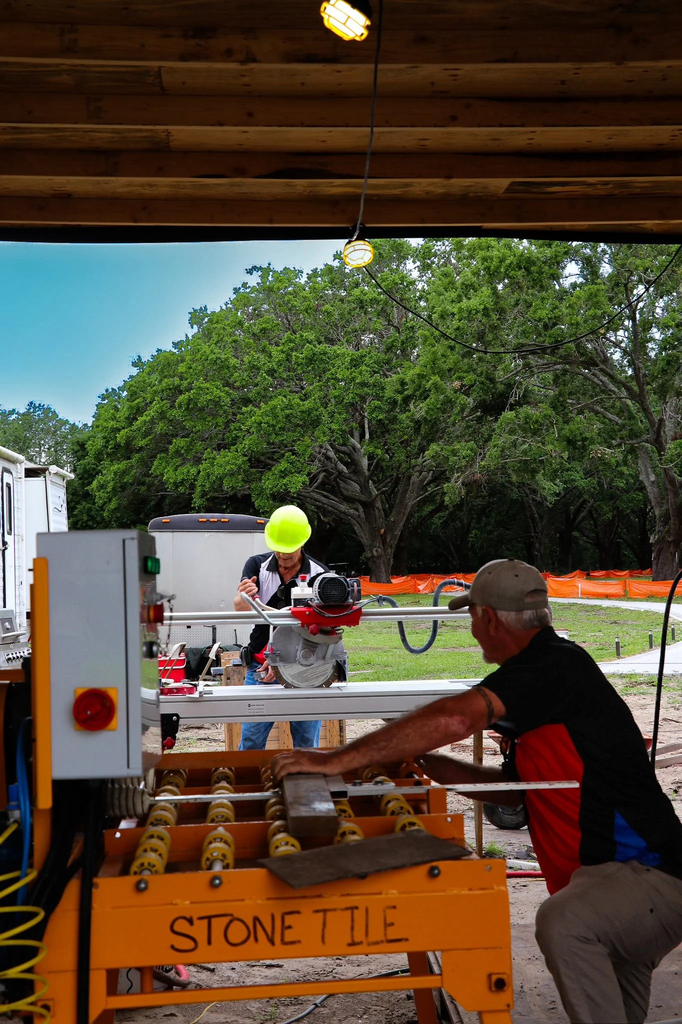 Two workers operating stone tile cutting equipment outdoors, with a large green tree and orange construction fencing in the background.