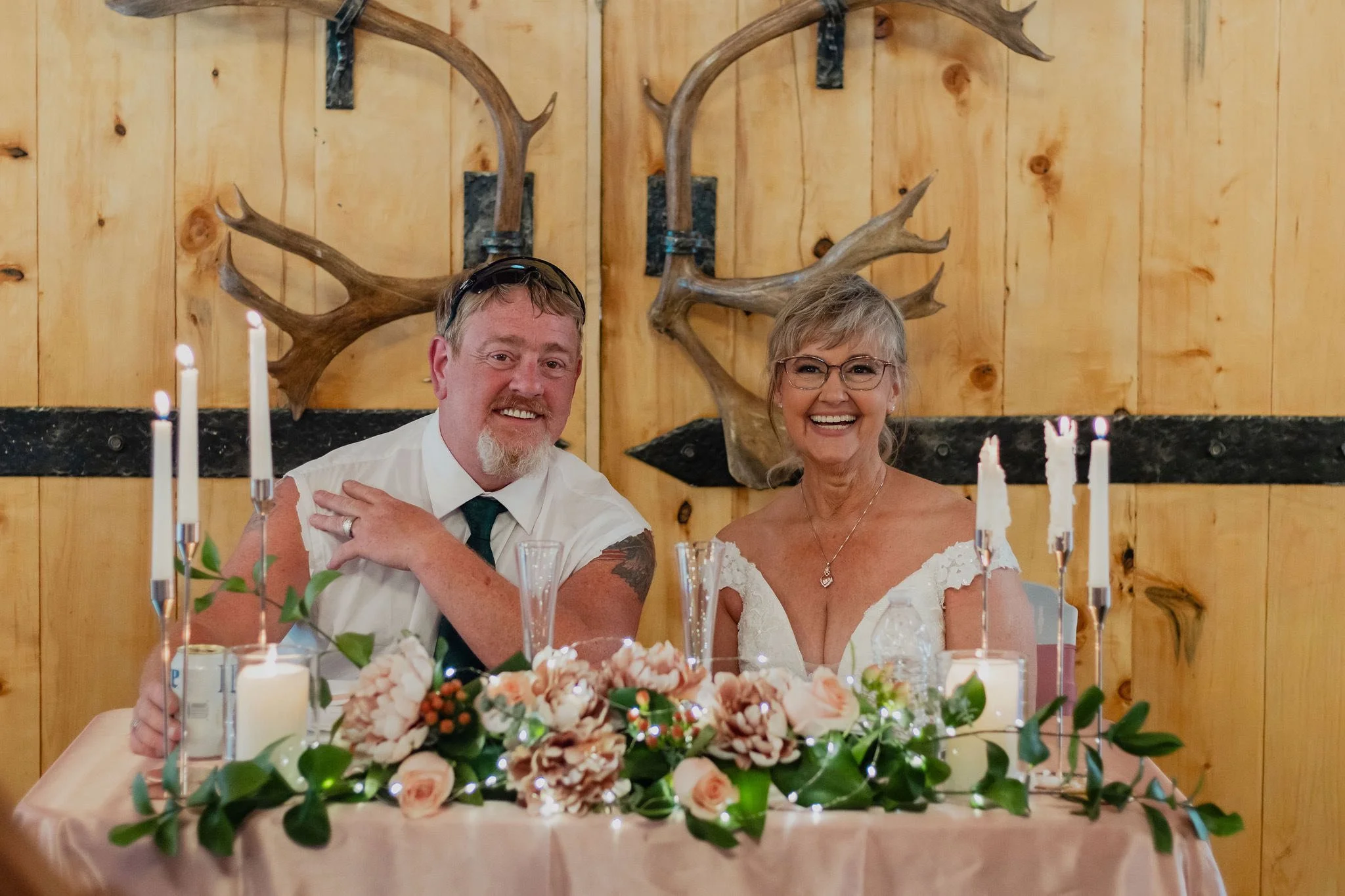 A Bride and Groom sit at a wedding head table during a wedding  with a wooden wall and mounted antlers in the background.