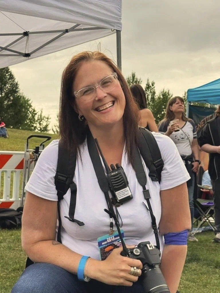A woman with brown hair, glasses, and a white t-shirt smiling at an outdoor event. She is carrying a camera and wearing a lanyard with event passes around her neck. She has a blue wristband and a blue armband on her arm.