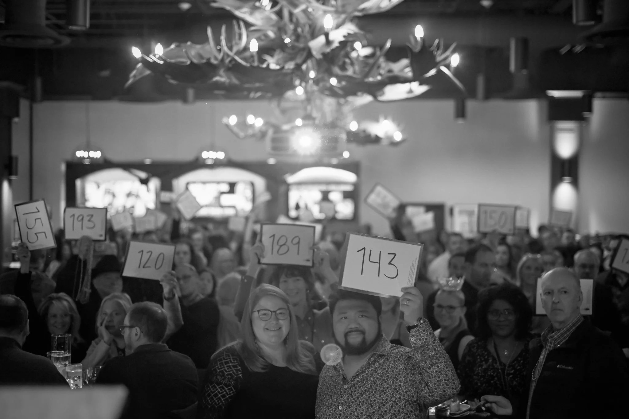 Crowd of people at an auction, holding up numbered paddles in a large room with chandeliers.