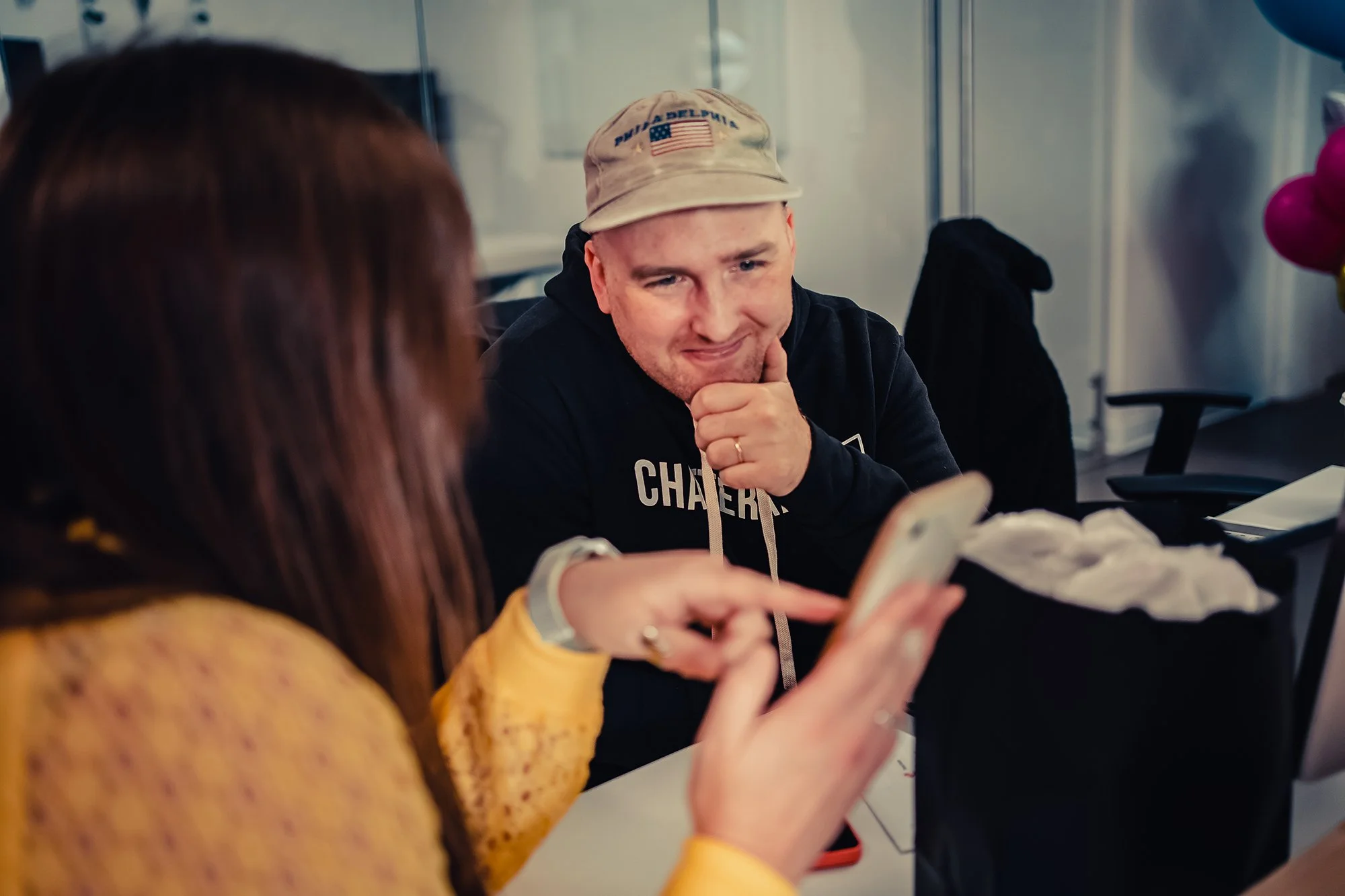 Two people sitting at a table, one is a woman with long brown hair wearing a yellow jacket and silver watch, pointing at her phone. The other is a man with a beige cap, black hoodie, with his hand on his chin, looking at the woman’s phone.