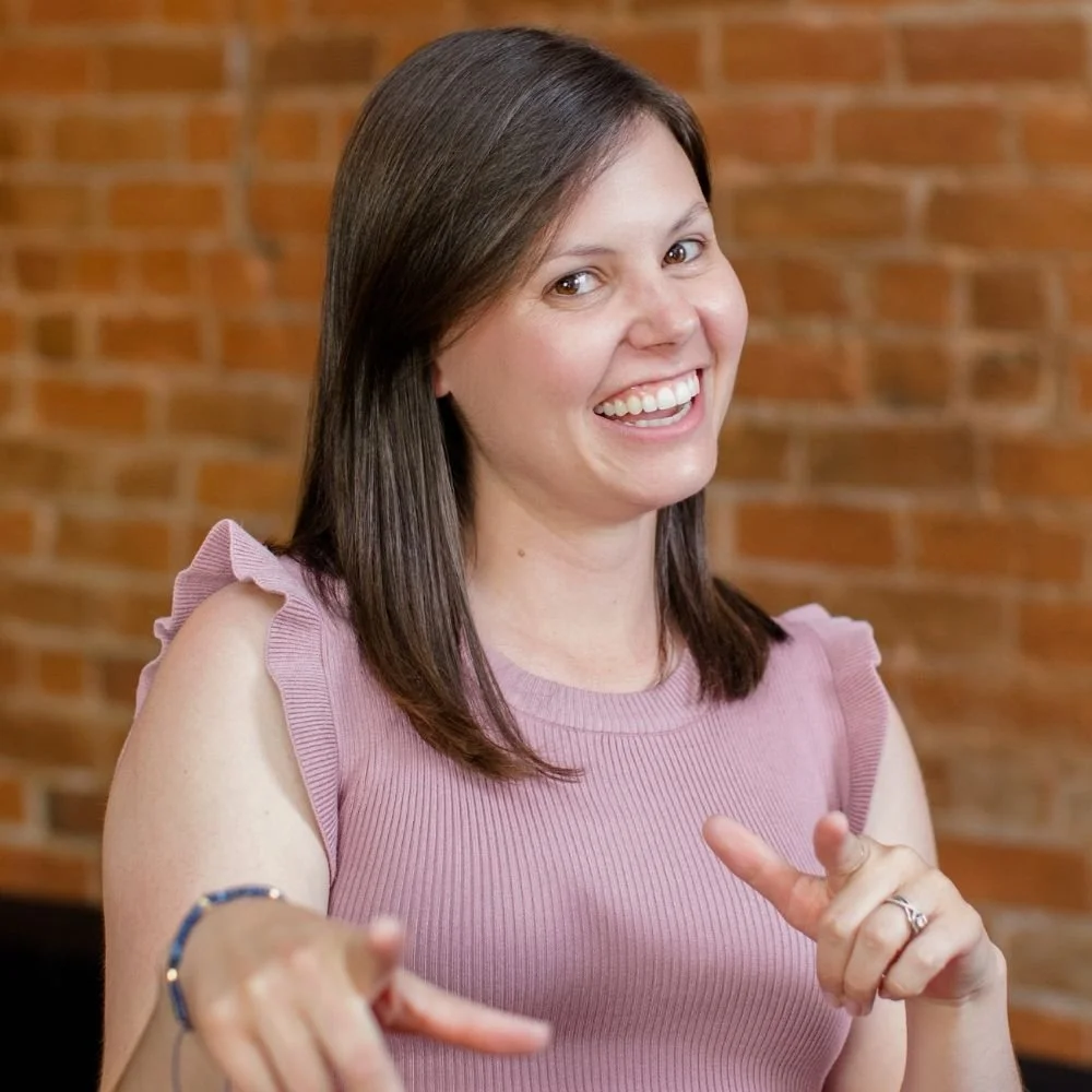 Smiling woman with shoulder-length dark hair wearing a pink sleeveless top with ruffled shoulders, pointing while standing in front of a brick wall.