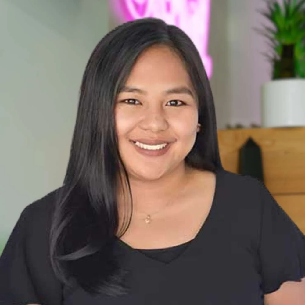 Smiling woman with long black hair wearing a black shirt, indoors with plants and a pink sign in the background.