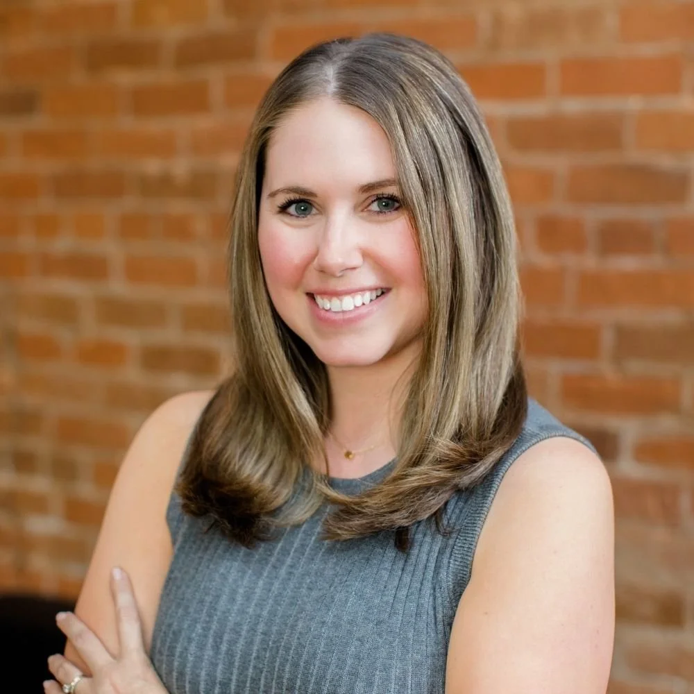 A woman with shoulder-length brown hair, smiling, wearing a sleeveless gray top, standing in front of a brick wall.