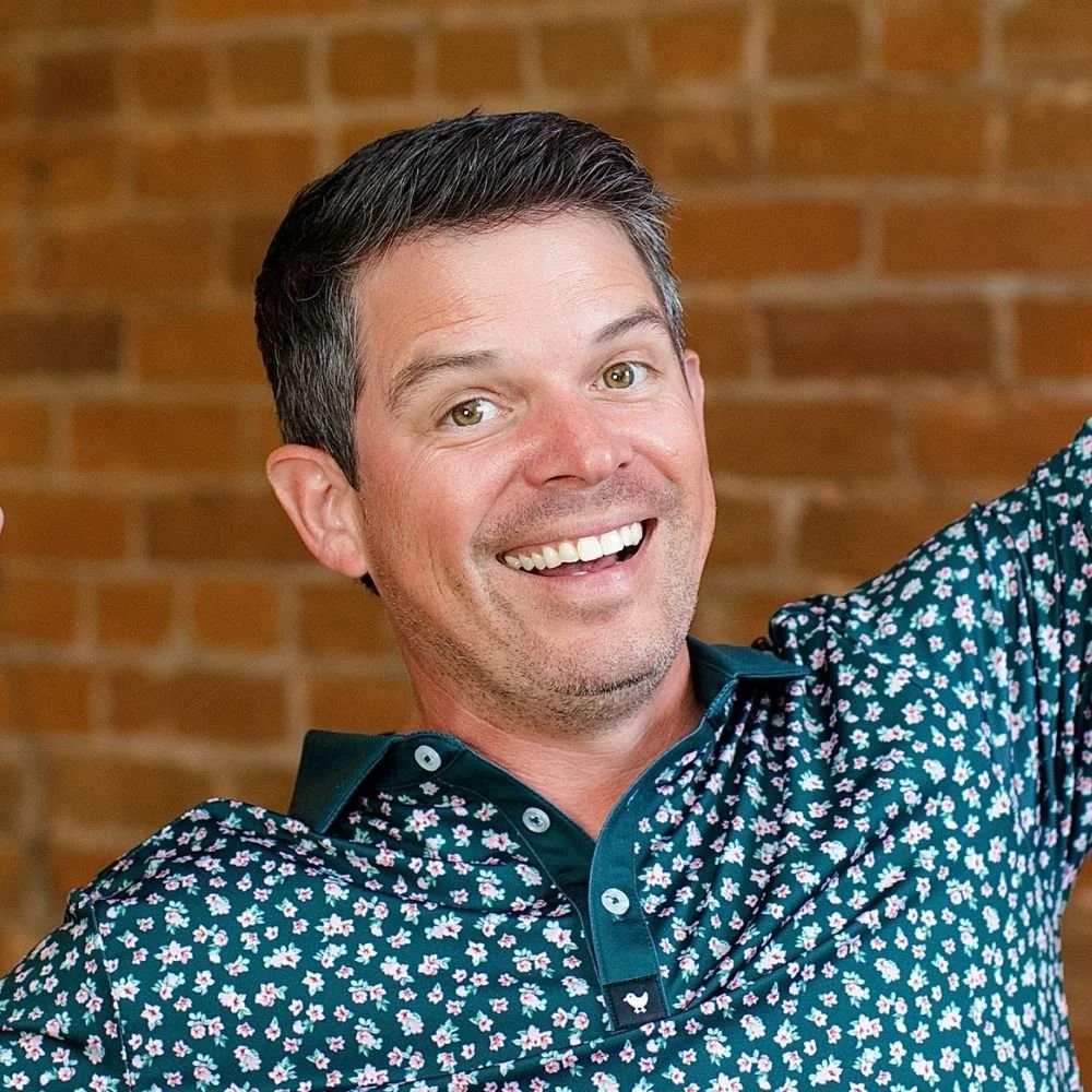 A man smiling, with short dark hair, wearing a dark floral shirt, in front of a brick wall.