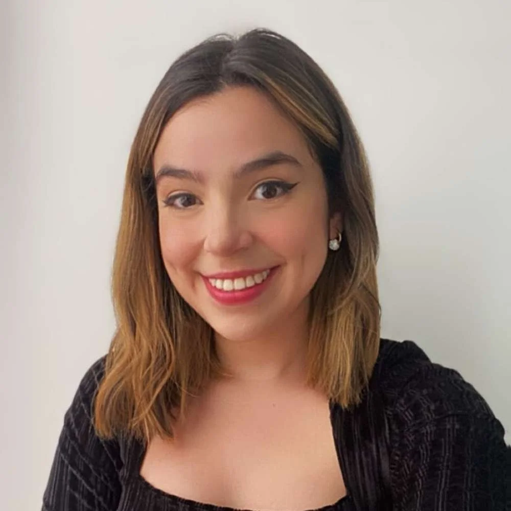 A smiling woman with shoulder-length light brown hair, wearing a black top, earrings, and makeup, against a plain white background.